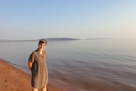 Mujer sonriente en la orilla de arena rojiza del lago Kariba, con el agua tranquila y el cielo despejado al fondo.