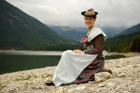 Frau in traditioneller Isarwinkler Tracht sitzt am Ufer eines Sees vor einer Bergkulisse.