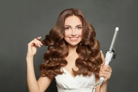 Uśmiechnięta kobieta z falowanymi brązowymi włosami, prezentująca lokówkę. Efektowne styling włosów.