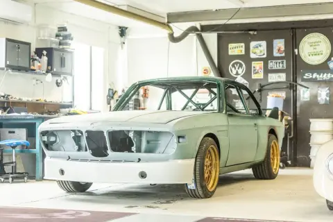En grön BMW E9 i verkstad, redo för renovering. Guldiga fälgar och en imponerande bur syns tydligt.
