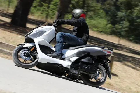 Persona conduce una SYM Jet X 125 blanca por una carretera arbolada. Las opiniones sobre la SYM Jet X 125 son positivas.