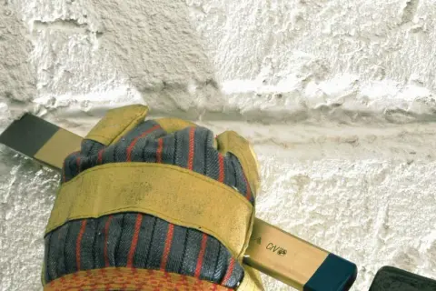 Mit einem Spachtel wird alter Putz entfernt. Die Arbeitshandschuhe schützen die Hände.