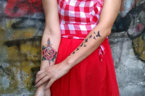 Un tatuaje de daga atraviesa una rosa roja en el brazo de una mujer. En el otro brazo, un grupo de pájaros vuelan.