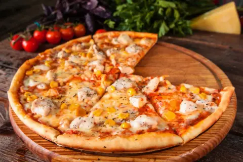 Pyszna pizza z kurczakiem i kukurydzą, podana na drewnianej desce, z pomidorkami koktajlowymi i bazylią w tle.