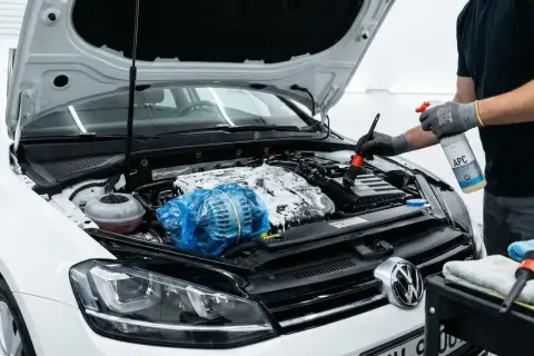 Persona con guantes rocía espuma y usa un cepillo para limpiar motor coche. El alternador está cubierto con plástico azul.