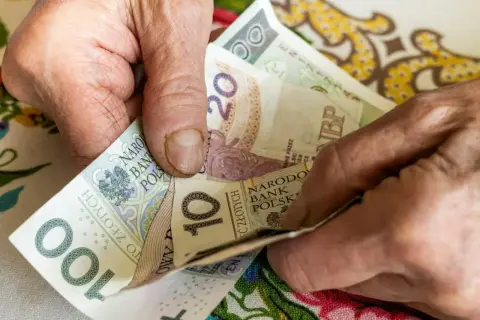 Dłonie starszej osoby trzymają polskie banknoty: 100, 20 i 10 złotych.