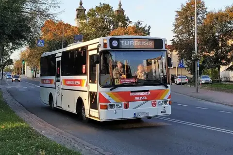 Biały autobus miejski z napisem "KOMUNIKACJA MIEJSKA" i "TURBUSS" jedzie ulicą. To część codziennych, busy przewozy po polsce.