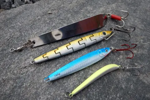 Vier Kunstköder für das Pilken vom Ufer liegen auf grauem Stein. Ein langer Silberpilker, ein gelb-silberner Wobbler, ein blauer Wobbler und ein gelber Gummifisch.