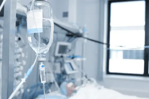 Kroplówka w szpitalnej sali, gdzie pacjent jest w spiaczce farmakologicznej. W tle aparatura medyczna i okno.