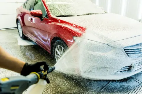 Czerwony samochód pokryty gęstą pianą aktywną podczas mycia. Ręka w czarnej rękawiczce trzyma pistolet do pianowania.