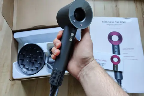 Dłoń trzymająca czarną suszarkę do włosów Dyson Supersonic obok pudełka i dyfuzora.