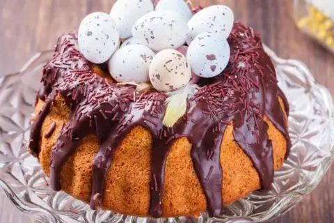Wilgotna babka gotowana, polana czekoladą i posypana czekoladową posypką, ozdobiona pisankami i piórkiem. Idealna na Wielkanoc.