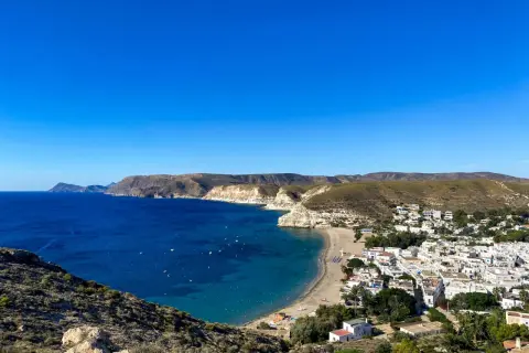 Vista panorámica de una **playa** con **agua amarga** de color azul intenso, un pueblo blanco y colinas áridas bajo un cielo despejado.