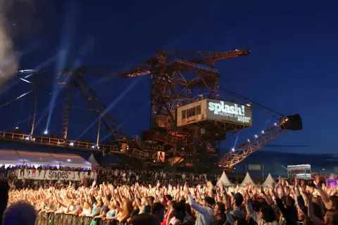 Große Menschenmenge jubelt bei einem Konzert vor einer riesigen Baggerschaufel mit dem Splash! Festival Line Up.