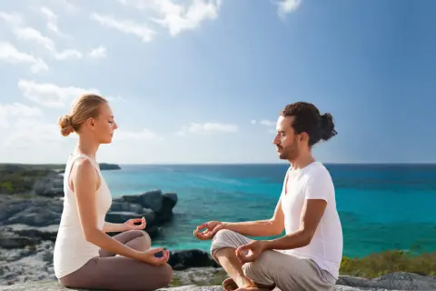Ein Paar meditiert in Lotusposition an einer felsigen Küste. Sie suchen nach spirituellen Ausbildungen, um ihre innere Ruhe zu finden.