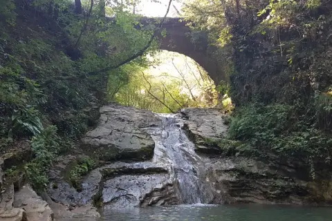 Cascate brescia: un piccolo salto d'acqua scende tra rocce e vegetazione lussureggiante, sotto un antico ponte ad arco.
