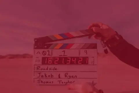 Dłoń trzyma klaps filmowy z zapisanymi informacjami o produkcji.