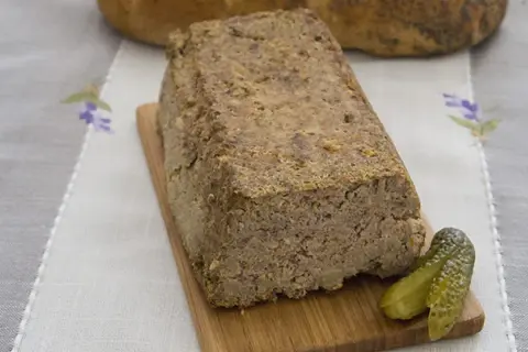 Domowy pasztet drobiowo-wieprzowy z wątróbką, podany z ogórkiem kiszonym na desce drewnianej.