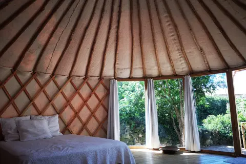 Intérieur d'une yourte gard, lit douillet sous la structure en bois, grandes fenêtres offrant une vue sur la verdure.