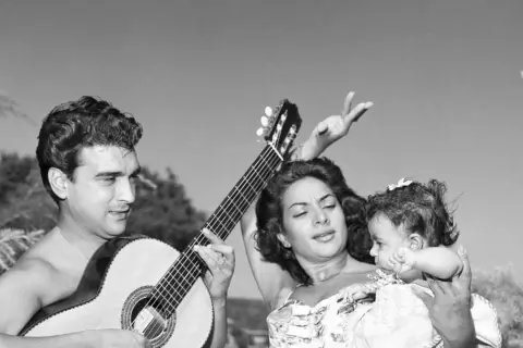 Hombre toca guitarra mientras mujer baila con bebé. Una escena de rumbas flamencas al aire libre.