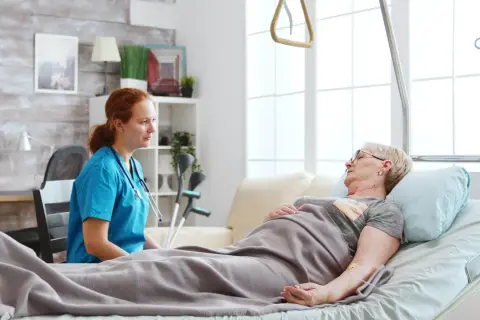 Pielęgniarka rozmawia z pacjentką leżącą w łóżku. W tle widać kule, co sugeruje, że pacjentka może potrzebować wsparcia w odżywianiu dojelitowym.