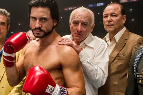 Roberto Duran, the legendary fighter, stands ready with red boxing gloves. His team, including Robert De Niro, offers support.
