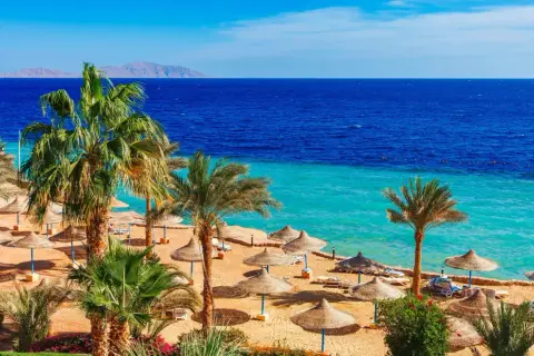 Solig strand med palmer och parasoller i Hurghada, Egypten. Blått hav och berg i horisonten.