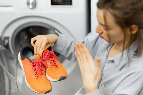 Donna con scarpe arancioni in mano, davanti a una lavatrice aperta. Scopri come lavare le scarpe in lavatrice per farle tornare come nuove.