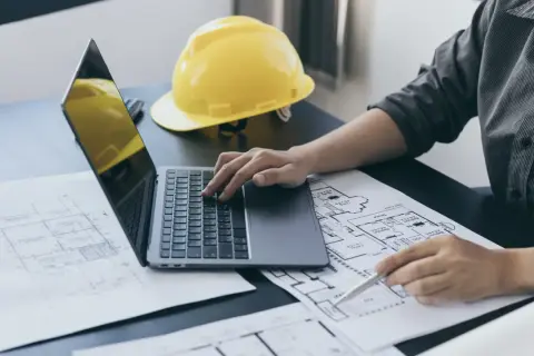 Architekt pracuje nad projektem, analizując plany i obliczając powierzchnię całkowitą budynku. Na biurku leży żółty kask.