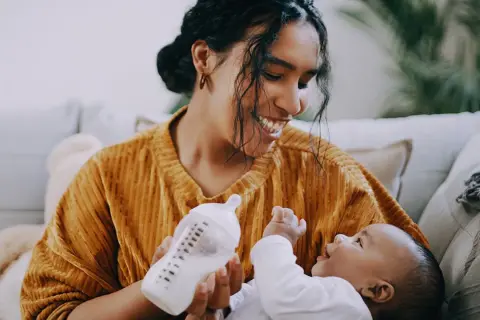 A mother smiles while paced feeding her newborn with a bottle. The baby looks up at her, reaching out a hand.