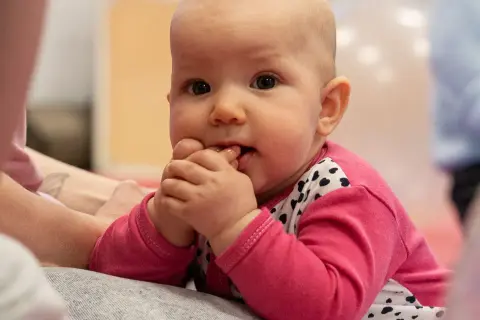 Malutkie dziecko w różowej bluzeczce z serduszkami, z paluszkami w buzi, delikatnie buja się w przód i tył, odkrywając świat.