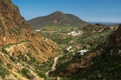 Paisaje árido con el **Cerro Negro** al fondo. Un camino serpentea entre barrancos y vegetación baja, llegando a unas casas blancas.