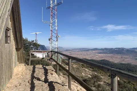 Vista panorámica desde la sierra del Carche, con una cabaña de madera, antena de comunicaciones y un paisaje de campos y montañas.