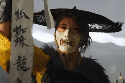 Postać w masce z Ghost of Tsushima, trzymająca zwój z japońskimi znakami. Osiągnięcie platyny w tej grze to wyzwanie!