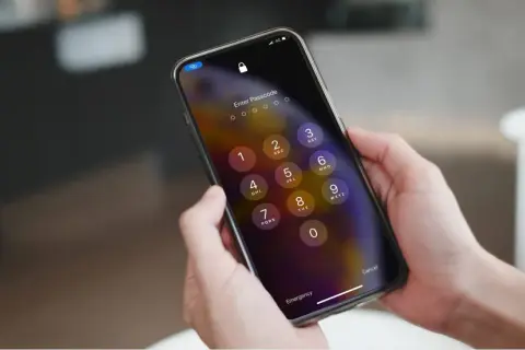 Dłonie trzymające iPhone'a z ekranem blokady i klawiaturą numeryczną.