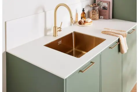 Évier en cuivre sous un robinet doré, sur un plan de travail stratifié blanc brillant. Des placards verts complètent l'ensemble.