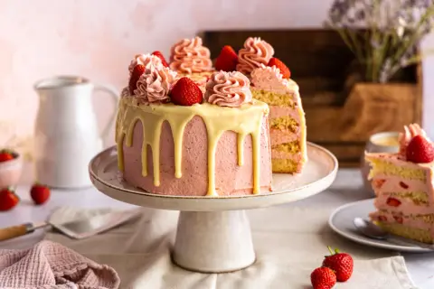 Eine wunderschöne Erdbeertorte, perfekt für jedes Erdbeertorte rezept. Mit rosa Buttercreme, weißen Schokoladenglasur-Tropfen und frischen Erdbeeren dekoriert.