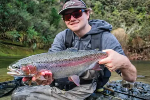 Glad fiske i Ånnaboda! En stolt fiskare håller upp en vacker regnbåge vid en forsande å.