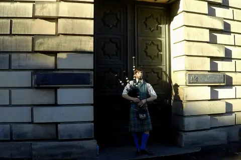 Mężczyzna w tradycyjnym szkockim stroju, grający na dudach. Ma na sobie kilt, kamizelkę i białą koszulę.