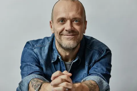 Uomo con barba corta, occhi azzurri, indossa una camicia di jeans. Le sue mani sono intrecciate, eccoti testo.