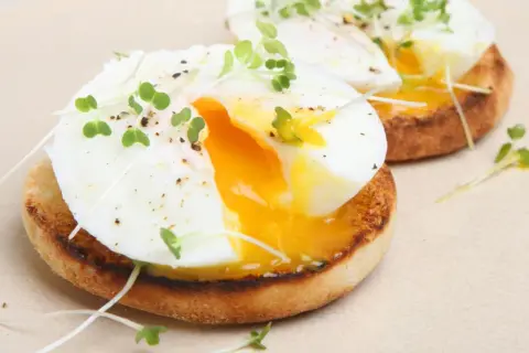 Dos deliciosos huevos poche sobre tostadas inglesas, adornados con brotes verdes y pimienta. La yema dorada fluye, invitando a un bocado perfecto.