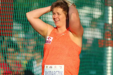 Ania Włodarczyk, po rzucie młotem rekord świata, z uśmiechem na twarzy i rękami na głowie, za zieloną siatką.