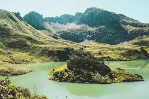 Grüne Hügel und ein türkisfarbener See laden zum Bergwandern in Bayern ein. Eine kleine Insel mit Bäumen liegt im Wasser.