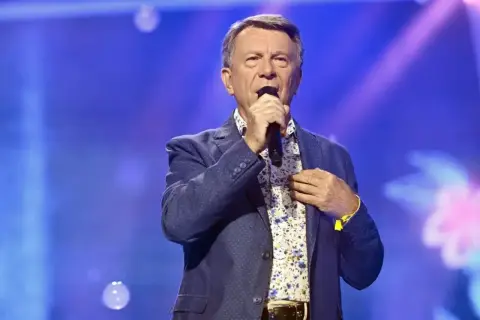 Wojciech Gąsowski w niebieskiej marynarce i kwiecistej koszuli śpiewa do mikrofonu na tle niebieskiej sceny.