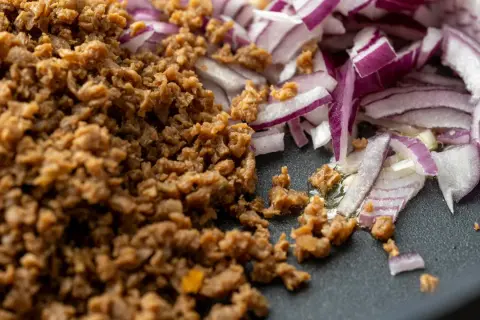 Gebratenes Hackfleischersatz-Granulat neben roten Zwiebeln in einer Pfanne.