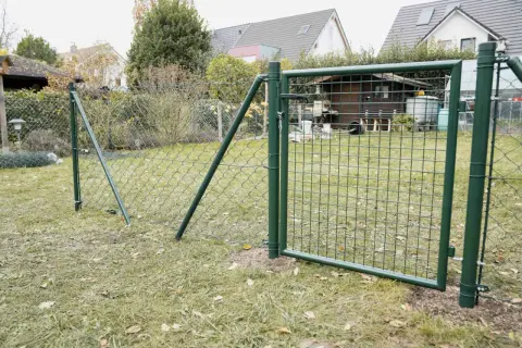 Ein grüner Maschendrahtzaun wird gespannt, mit einem Tor, das offen steht. Im Hintergrund ein Garten mit Häusern und einem Schuppen.