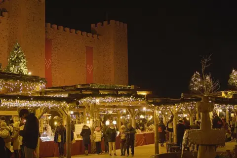 Mercado navideño bajo un castillo iluminado. Gente pasea entre puestos decorados con luces, mostrando como se celebra la navidad.