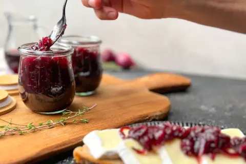Eine Hand schöpft mit einem Löffel köstliche zwiebelmarmelade einfach aus einem Glas. Dazu passt Brot mit Käse.