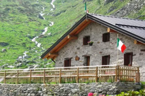 Rifugio in pietra con bandiera italiana, circondato da prati verdi e un ruscello. Un luogo ideale per rifugi val camonica facili.