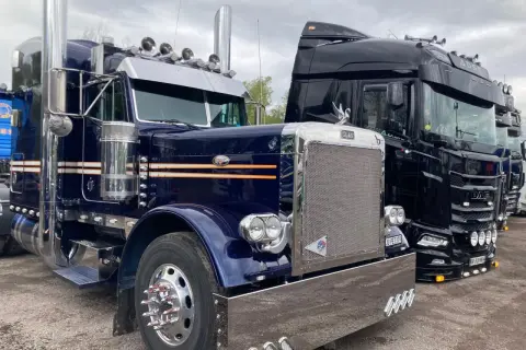 Deux camions américains, un bleu et un noir, brillent sous un ciel couvert. Le bleu, un Peterbilt, arbore des touches orange et beaucoup de chrome.
